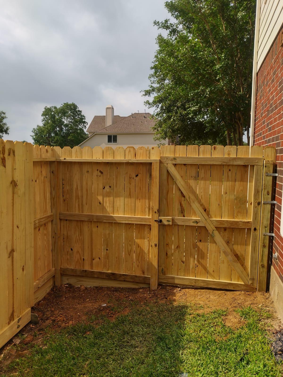 New pressure-treated pine privacy fence turning a corner beside a brick house, with a single walk gate braced by a diagonal board and hung on hinges at the house end