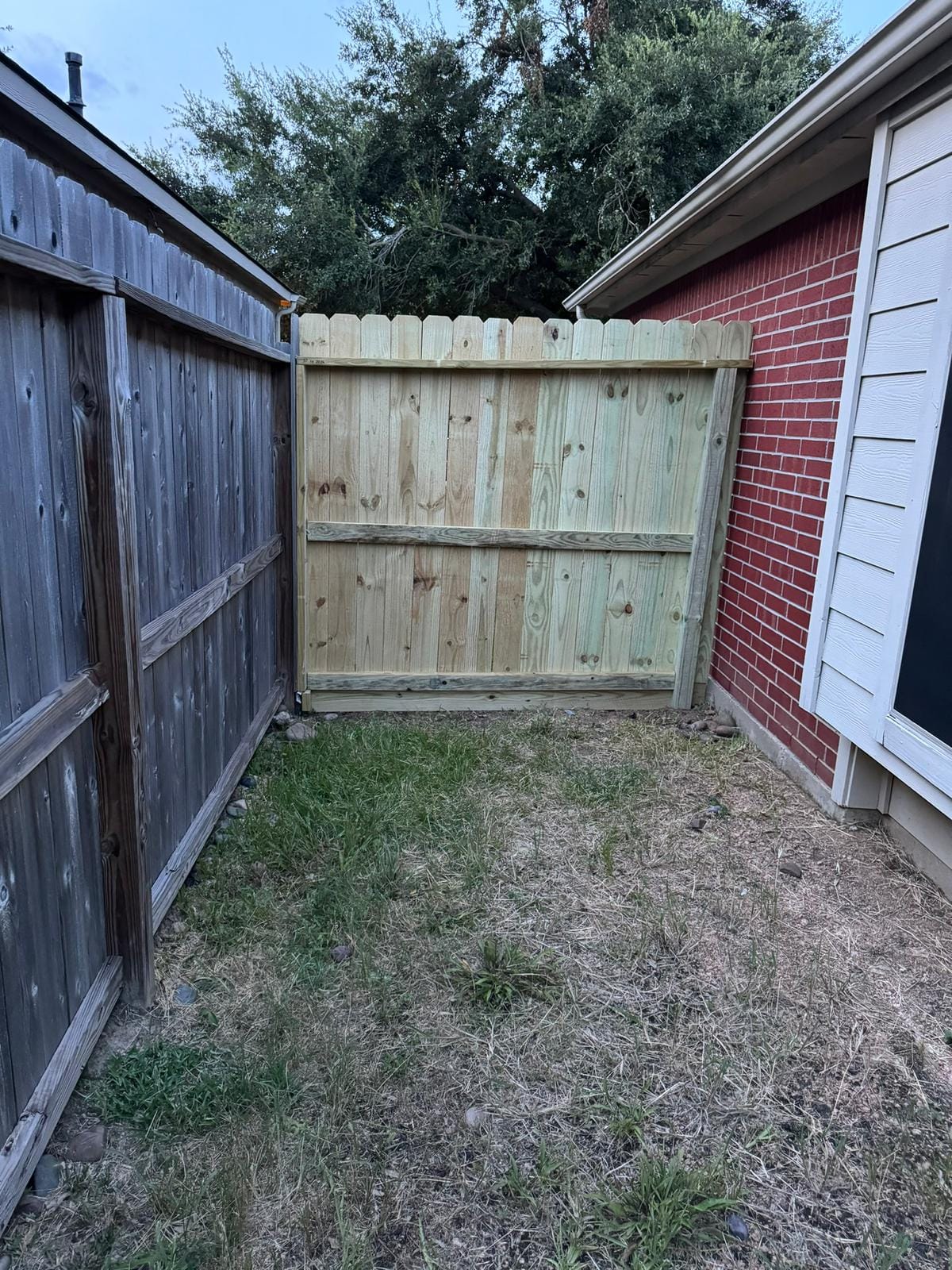 New light-colored wood fence panel meeting an older weathered fence at a side yard beside a red house