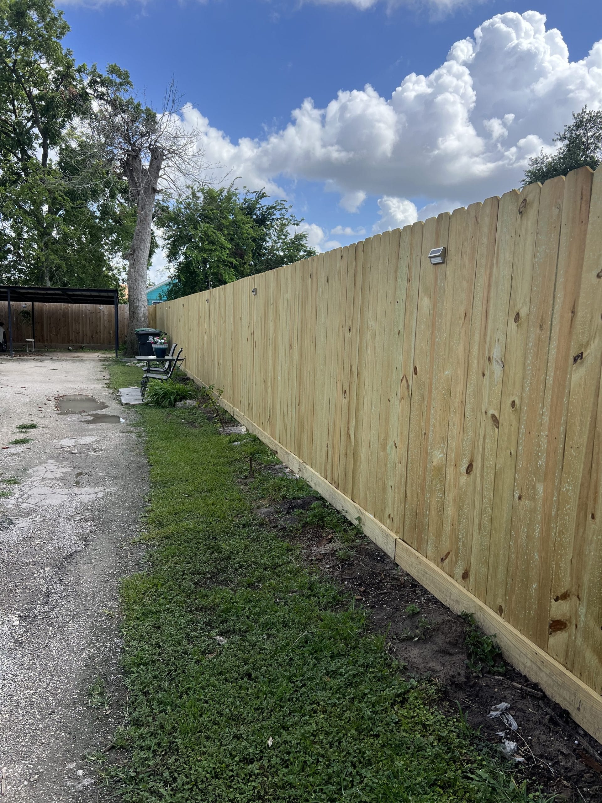 New pressure-treated pine privacy fence with a bottom rot board along a gravel drive