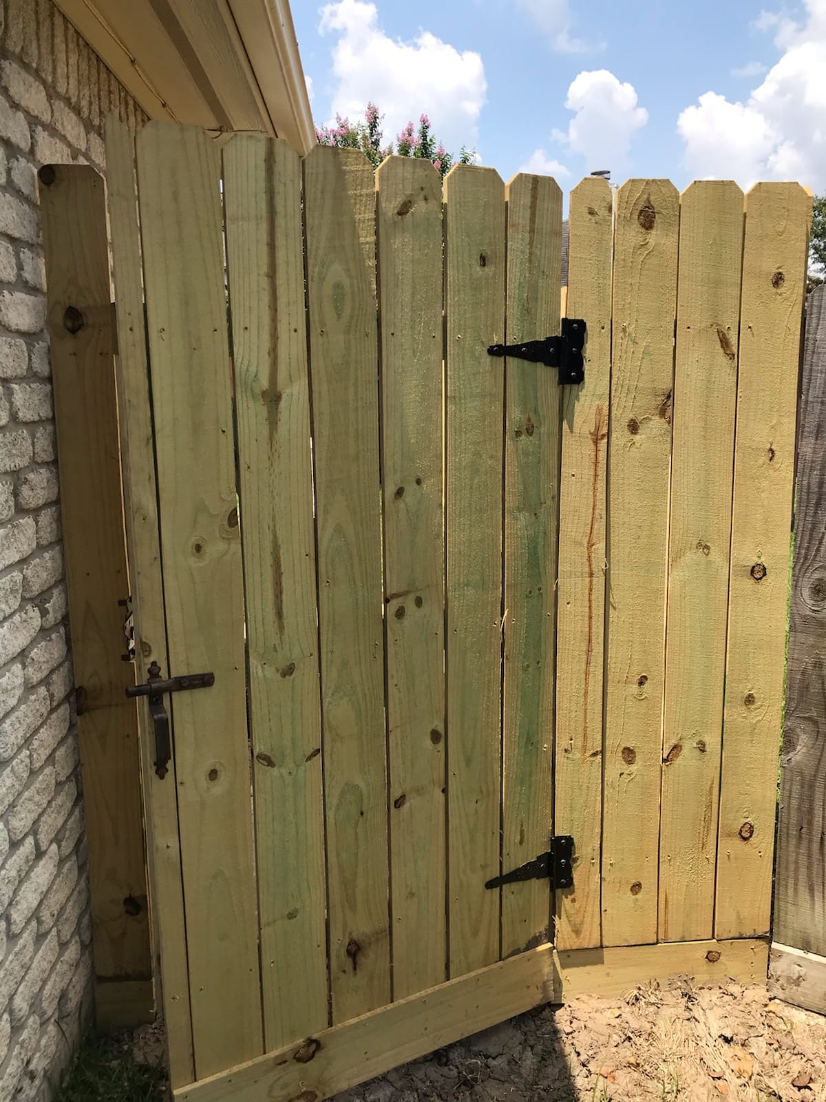 Wood privacy gate on black strap hinges beside a brick wall, its bottom board just above loose soil