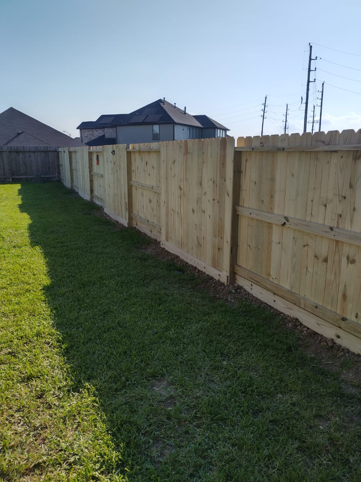 New dog-ear pine privacy fence running along the edge of a mown lawn, seen from its rail side, with neighboring two-storey houses beyond