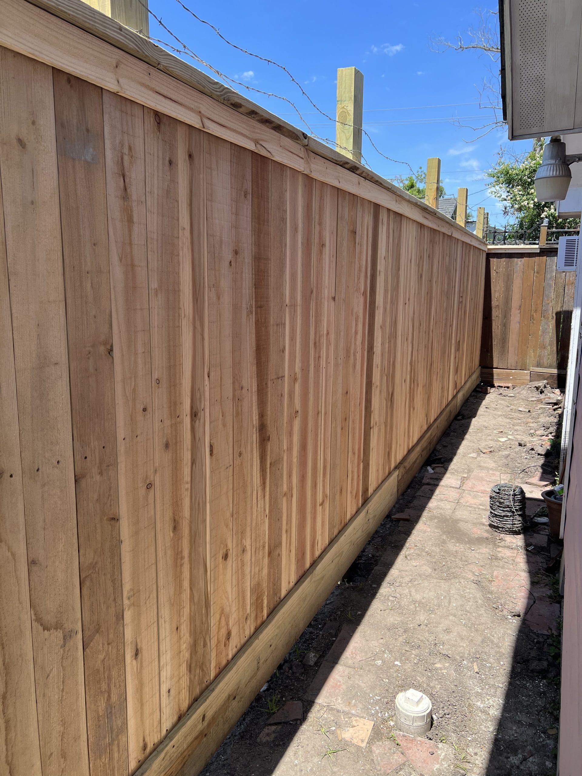 Tall new wood privacy fence with a top cap, a trim board and a bottom rot board running along a concrete side-yard walkway, with the smooth board face toward the walk
