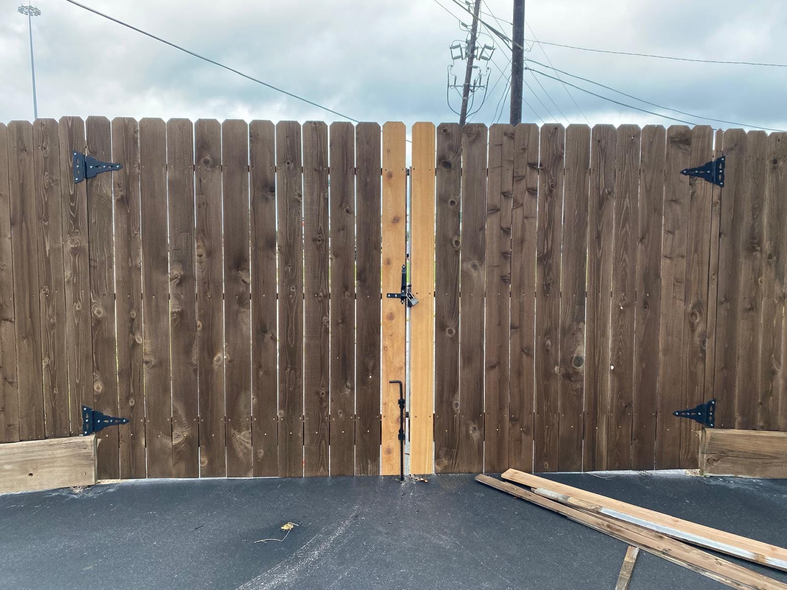 Stained wood double drive gate with black strap hinges, closed across an asphalt lot, with a vertical drop rod at the centre where the two leaves meet