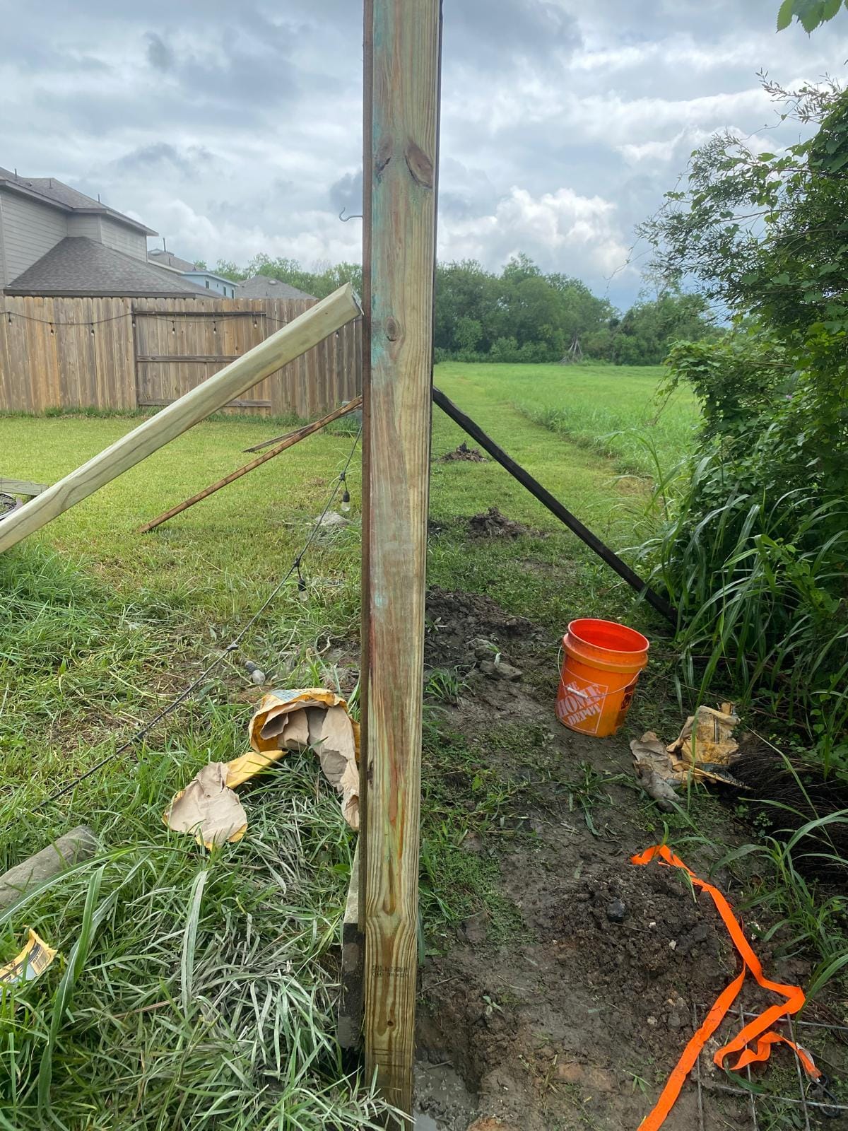 A braced wood corner post at the edge of an open grassy field, with an orange strap and bucket on the ground and an older wood fence and houses in the distance