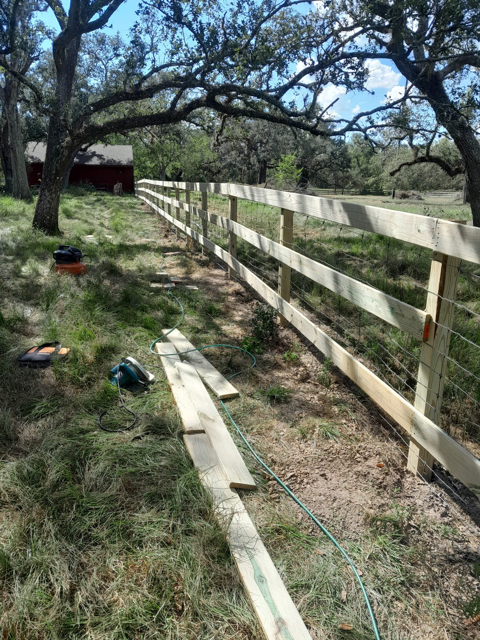 Pine three-rail fence with field wire behind the rails running under large oak trees, with lumber and a circular saw on the grass during construction