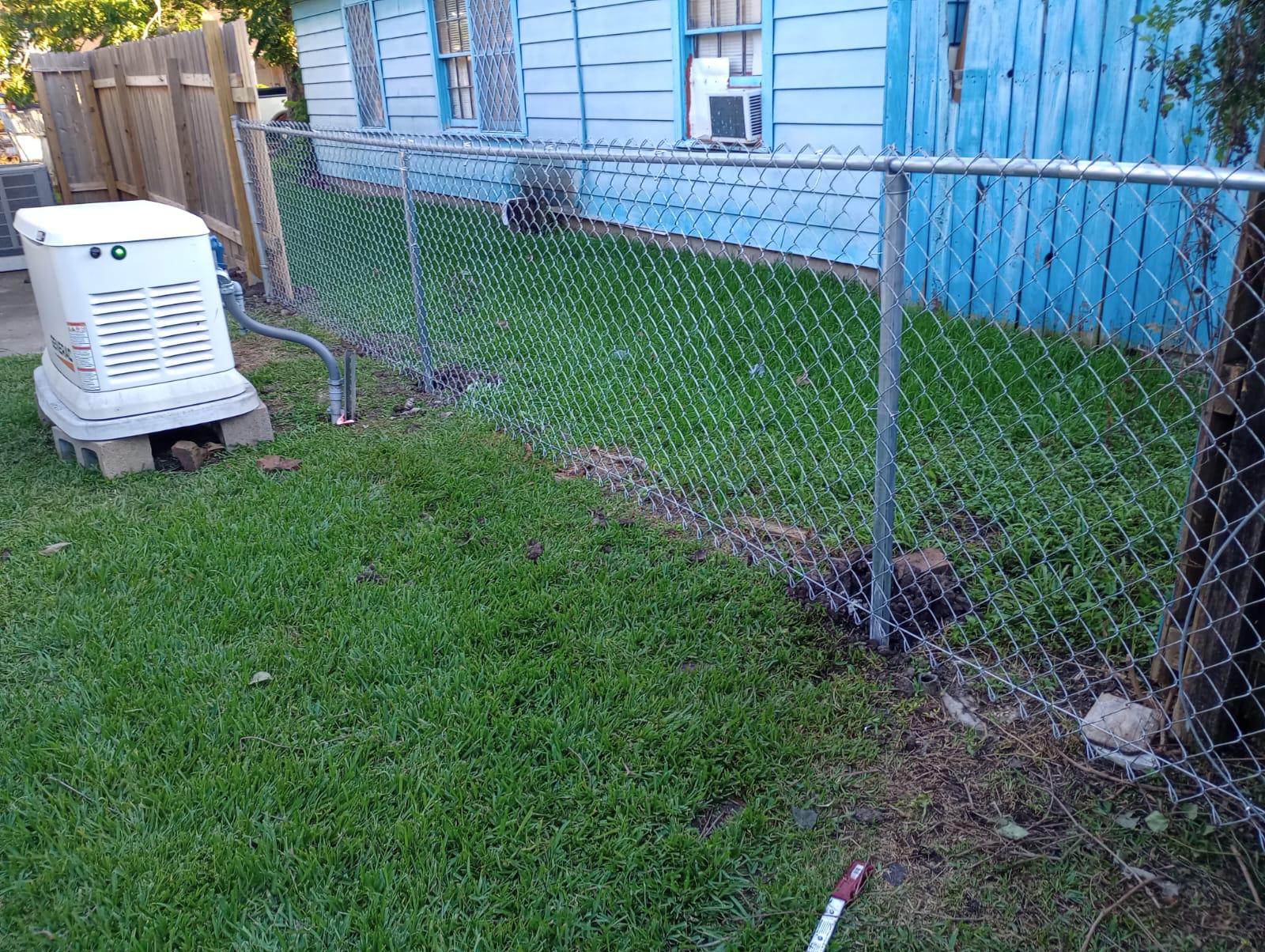 Standby generator set on concrete blocks in a side yard, beside a new galvanized chain-link fence and a blue house