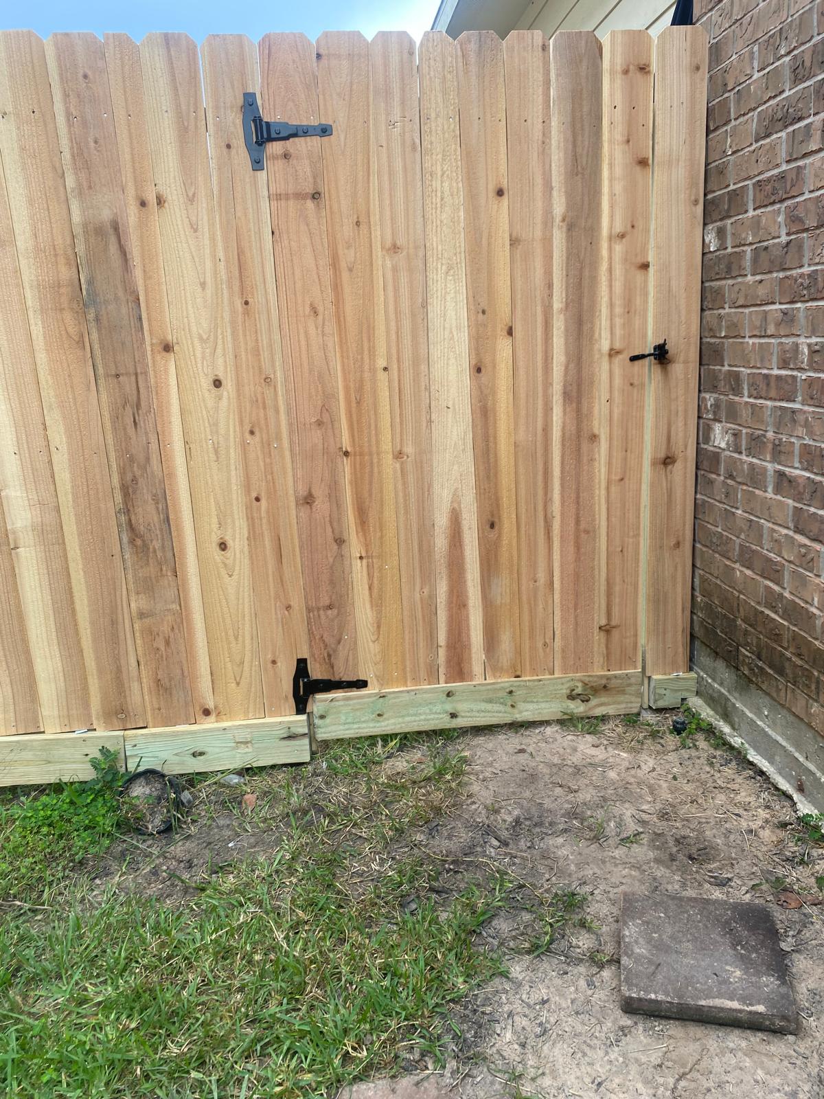 Wood privacy fence and gate with black hardware beside a brick house, with a bottom board along the grass